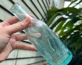 May include: A vintage, clear glass bottle with a light blue tint. The bottle has embossed text that reads "AMERICAN BOTTLING CO. SARA USA". The bottle is held against a background of green foliage and a weathered wooden surface.