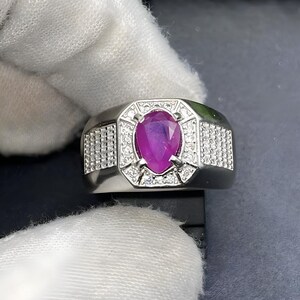 Könnte beinhalten: Ein silberner Ring mit einem großen, oval geschliffenen Rubin im Zentrum. Der Ring ist mit kleinen, klaren Edelsteinen besetzt und hat ein rechteckiges Design. Der Ring wird vor einem dunklen Hintergrund gezeigt.