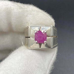 May include: A silver ring with a square design, featuring a vibrant, round, pink gemstone at the center. The ring is held against a dark background, showcasing the gemstone's color and the ring's details.