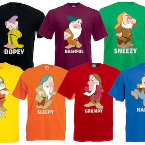 Könnte beinhalten: Eine Sammlung von sieben farbigen T-Shirts, die jeweils einen anderen Zwerg-Charakter aus Schneewittchen zeigen. Die Shirts sind schwarz, weinrot, grün, gelb, orange, rot und blau. Jedes Shirt hat den Namen des Charakters darunter.
