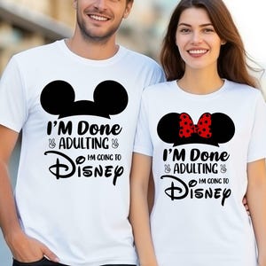 Op de afbeelding: Twee witte T-shirts met zwarte Mickey Mouse oren en de tekst "I'm Done Adulting I'm Going To Disney" met vredestekens. Het shirt van de vrouw heeft een rode strik op de oren.