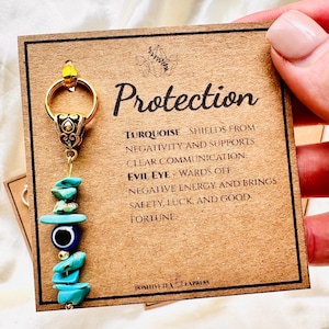 May include: A gold-coloured keychain with a charm featuring turquoise stones and an evil eye bead. The card reads "Protection" with details about the charm's properties. The keychain is held by a person.