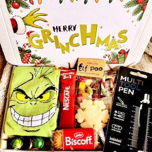May include: A gift box labelled Merry Grinchmas containing a green sock with a Grinch face, a red Nescafe 3-in-1 packet, a bag of Elf Poo candy, a snowflake cookie, a Lotus Biscoff biscuit, and a black multi-tool pen with a visible ruler scale.