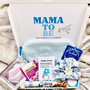 May include: A gift box labeled "MAMA TO BE" with a stork illustration. Inside are a sleep mask, tea, lip mask, scrunchie, keychain, and a Galaxy chocolate bar. The box is on a soft, textured surface.