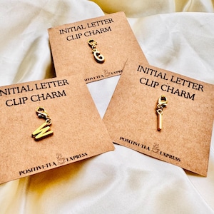 May include: Three gold initial letter clip charms on brown cards. Each card reads "INITIAL LETTER CLIP CHARM" and has the "POSITIVI-TEA EXPRESS" logo. The letters are "W", "E", and "I".