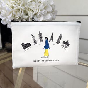 May include: White canvas travel pouch with a black zipper. The pouch features a black and white illustration of a person walking towards famous landmarks, including the Statue of Liberty and the Eiffel Tower. Text on the pouch reads "look at the world with love."