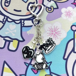May include: A silver-toned metal keychain featuring a heart-shaped clasp and three dangling charms. The charms include a black and white heart, a white bear-like figure with black outlines, and a black and white text charm reading TRASH.