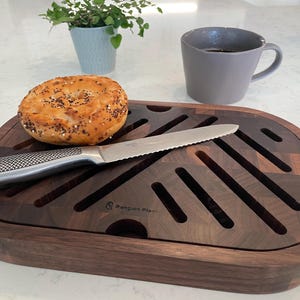May include: A dark wood breadboard with a bagel and serrated knife. The board has a grid pattern and a rounded shape. A gray mug and small potted plant are in the background.