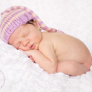 May include: A newborn baby sleeping soundly on a white blanket, wearing a pink and white striped knitted hat with a long pointy tip.