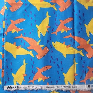May include: Blue fabric with a repeating pattern of orange and yellow cartoon sharks and small purple fish. The fabric has text at the bottom that reads "Beach", "David Walker for Free Spirit", and "Westminster Fibers # PWOW074 Sharks".