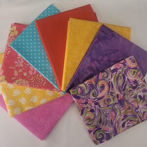 May include: A collection of folded fabric squares in various colors and patterns, including a teal polka dot print, a solid orange, a yellow floral, a purple batik, a pink floral, a yellow teacup print, a solid pink, and a purple swirl pattern.
