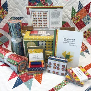 May include: A quilting kit featuring a yellow tin, fabric rolls, and squares with floral and geometric patterns. Includes a pattern card titled Brio, a card with the quote Be yourself, everyone else is already taken, and two spools of thread.