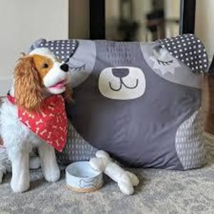 May include: A gray and white dog-shaped pillow with a white bone toy and a white bowl with a bone design. The pillow has a black nose, white teeth, and closed eyes with eyelashes.