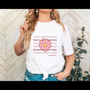 May include: White t-shirt with a pink flower graphic and the text "Pray Like Hannah" repeated in a pattern. The text "1 Samuel 1:11" is also included.