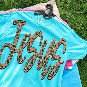May include: A turquoise t-shirt featuring the word "Jesus" in a large, decorative font. The letters are filled with a leopard print design and outlined in brown. The shirt is hanging on a black clothes hanger.
