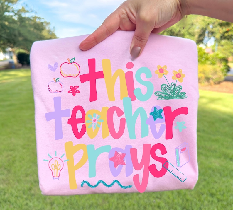 Op de afbeelding: Een lichtroze t-shirt met de tekst "This Teacher Prays" in kleurrijke letters. Het ontwerp bevat illustraties van appels, bloemen, sterren en een gloeilamp. Een vrolijk ontwerp, geschikt voor leraren.