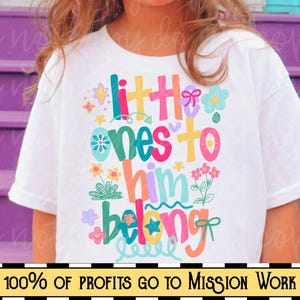 Op de afbeelding: Wit t-shirt met de tekst "little ones to him belong" in kleurrijke, speelse letters, versierd met bloemen en sterren. De onderkant van het shirt heeft een banner met de tekst "100% OF PROFITS GO TO MISSION WORK".