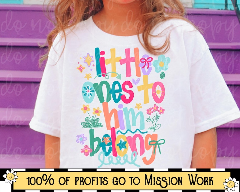 Op de afbeelding: Wit t-shirt met de tekst "little ones to him belong" in kleurrijke, speelse letters, versierd met bloemen en sterren. De onderkant van het shirt heeft een banner met de tekst "100% OF PROFITS GO TO MISSION WORK".