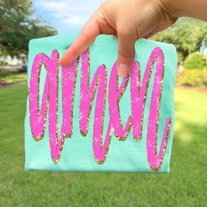 Op de afbeelding: Een lichtblauwgroen t-shirt met het woord "WHIM" in felroze, omlijnd met gouden glitter. Het shirt wordt omhooggehouden tegen een achtergrond van groen gras en bomen.