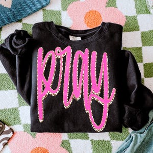 Puede incluir: Sudadera negra con la palabra "PRAY" en letras grandes de color rosa brillante con contorno de estampado de leopardo. La sudadera está doblada sobre una superficie colorida y estampada. Las mangas están dobladas.