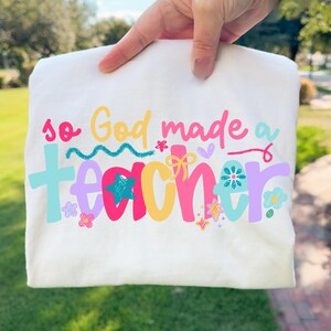 Op de afbeelding: Wit T-shirt met de kleurrijke tekst "so God made a TEACHER". De tekst is in verschillende kleuren, waaronder roze, blauw, geel en paars, met bloemen- en steraccenten. Het shirt wordt door een hand omhoog gehouden.