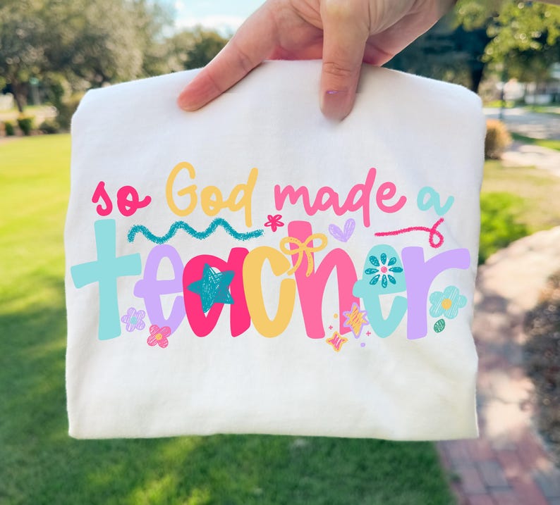 Op de afbeelding: Wit T-shirt met de kleurrijke tekst "so God made a TEACHER". De tekst is in verschillende kleuren, waaronder roze, blauw, geel en paars, met bloemen- en steraccenten. Het shirt wordt door een hand omhoog gehouden.