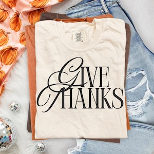 May include: A cream-colored short-sleeve t-shirt with the black text GIVE THANKS printed in a stylized serif font on the front. The shirt features a Comfort Colors brand label at the inner neckline and is layered over other garments.