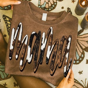 May include: A brown sweatshirt with the word "MAMA" in a bold, cursive font. The letters have a cow print design with white, brown, and black spots, outlined in black. The sweatshirt is held by hands, and the background includes candles and a patterned cloth.