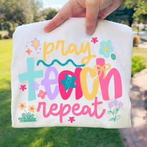 Op de afbeelding: Een gevouwen wit t-shirt met de woorden "pray teach repeat" in een kleurrijk, speels lettertype. Het ontwerp bevat sterren, bloemen en een kruis, allemaal in heldere kleuren. Het shirt wordt door een hand omhoog gehouden.
