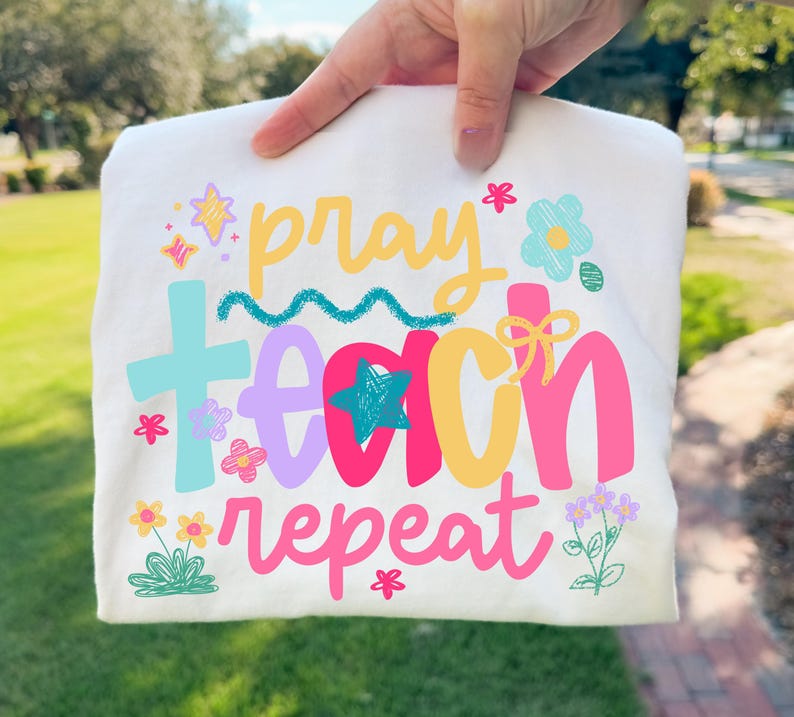 Op de afbeelding: Een gevouwen wit t-shirt met de woorden "pray teach repeat" in een kleurrijk, speels lettertype. Het ontwerp bevat sterren, bloemen en een kruis, allemaal in heldere kleuren. Het shirt wordt door een hand omhoog gehouden.