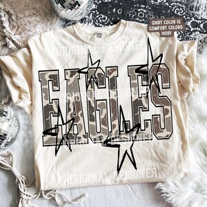 Peut inclure: Un t-shirt à manches courtes ivoire avec le mot EAGLES en grandes lettres majuscules arborant un motif camouflage marron et beige. Le design inclut des étoiles aux contours noirs superposées sur le texte. Une étiquette indique SHIRT COLOR IS COMFORT COLORS IVORY.