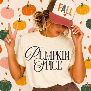 May include: A cream-colored t-shirt with the black text PUMPKIN SPICE printed on the front. The shirt is paired with a brown and cream trucker hat featuring the word FALL in colorful letters and a small heart icon on the front panel.