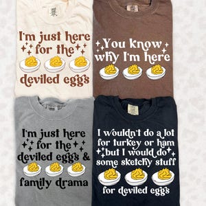 May include: Four crew neck t-shirts in cream, brown, gray, and black. Each shirt features a graphic of three deviled eggs and text. The cream shirt says I'm just here for the deviled eggs, brown says You know why I'm here, gray says I'm just here for the deviled eggs & family drama, and black says I wouldn't do a lot for turkey or ham but I would do some sketchy stuff for deviled eggs.