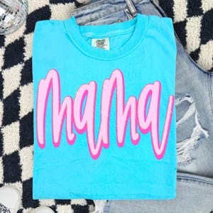 Op de afbeelding: Een fel turquoise t-shirt met korte mouwen waarop het woord mama is gedrukt in een gestileerd roze lettertype met een lichtroze omlijning. Het shirt heeft een ronde hals en een Comfort Colors merklabel zichtbaar bij de kraag.