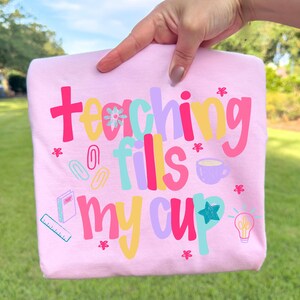 Op de afbeelding: Lichtroze t-shirt met de kleurrijke tekst "teaching fills my cup". Het ontwerp bevat een koffiekopje, een gloeilamp en schoolbenodigdheden.