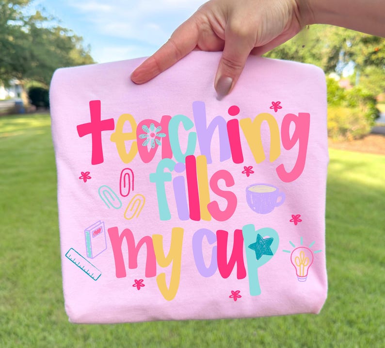 Op de afbeelding: Lichtroze t-shirt met de kleurrijke tekst "teaching fills my cup". Het ontwerp bevat een koffiekopje, een gloeilamp en schoolbenodigdheden.