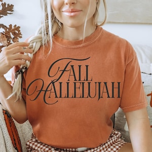 May include: A burnt orange short-sleeve t-shirt featuring the black text "FALL HALLELUJAH" printed across the chest in a stylised serif font. The garment has a crew neckline and a relaxed fit, showcasing a solid, muted colour throughout the fabric.