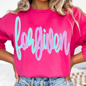 May include: A bright pink sweatshirt with the word "forgiven" in a script font. The word is outlined in light blue and purple. The sweatshirt is a solid color and has long sleeves.