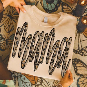 May include: A beige sweatshirt featuring the word "Mama" in a large, decorative font. The letters have a leopard print pattern with black outlines. The sweatshirt is held by hands, and the background has a butterfly design.