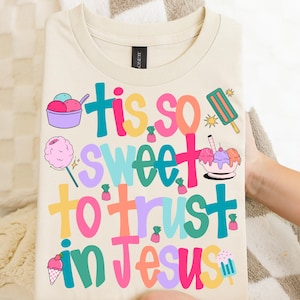 May include: Cream-colored t-shirt with the phrase "Tis so sweet to trust in Jesus" in colorful, playful lettering. The design includes ice cream, cotton candy, and other sweet treats.