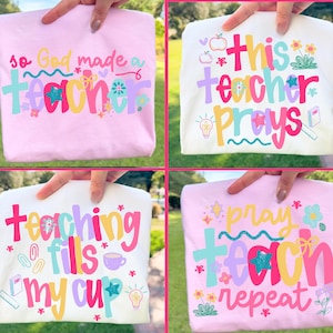 Op de afbeelding: Vier gevouwen t-shirts in roze en wit, elk met een kleurrijk tekstontwerp. De ontwerpen bevatten zinnen als "so God made a teacher", "this teacher prays", "teaching fills my cup" en "pray teach repeat".