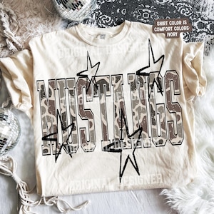 May include: An ivory t-shirt featuring the word MUSTANGS in large block letters with a brown and white cow print pattern. The design includes black outlined star shapes positioned around the text. A small tag indicates the shirt color is ivory.