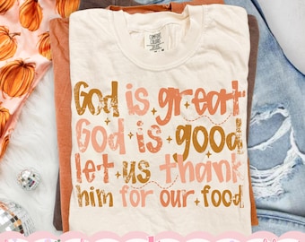 May include: A cream-coloured t-shirt featuring the text "God is great God is good let us thank him for our food" in a distressed, multi-coloured font of orange, pink, and brown, with small star and dashed line accents.