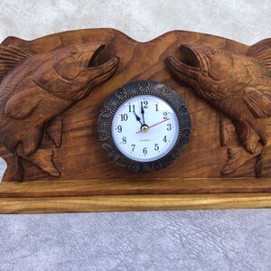 May include: A wooden clock with two carved fish on either side of the clock face. The clock face is white with black numbers and hands. The clock is on a wooden base.