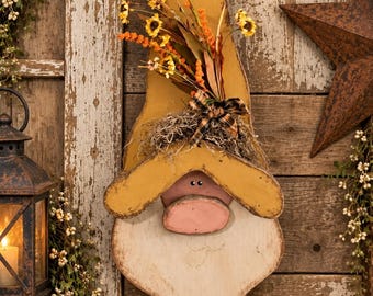 May include: A wooden wall hanging shaped like a gnome with a tall mustard yellow hat, a pink nose, and a white beard. The hat is decorated with a cluster of artificial orange and yellow flowers, a small plaid bow, and dried natural fibers.