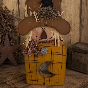 May include: A wooden scarecrow figure featuring a brown hat with straw accents, a painted face, and a plaid scarf. The body is a yellow rectangular block with a dark plaid pattern, a single gray button, and a crescent moon cutout.