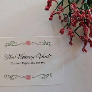May include: A white business card with the text "The Vintage Vault" and "Curated Especially For You" in elegant script. The card is decorated with floral accents and is accompanied by a sprig of red berries.