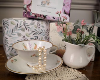 May include: A white porcelain teacup and saucer with gold rims and floral accents, a string of faux pearls, a small white ceramic creamer with artificial flowers, a silver spoon, and a crocheted doily, set before boxes with floral patterns.