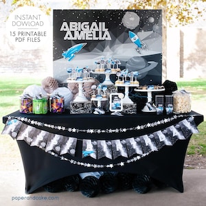 Puede incluir: Una mesa de fiesta de temática espacial en blanco y negro con una pancarta que dice "Feliz cumpleaños". La mesa está decorada con una nave espacial, planetas y estrellas. También hay varios frascos de dulces y palomitas de maíz en la mesa. El telón de fondo es un póster de temática espacial en blanco y negro con los nombres "Abigail Amelia" impresos en él.