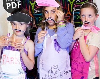 Puede incluir: Tres niños sostienen accesorios de cabina de fotos frente a un fondo de Torres Eiffel. Los accesorios son una boina negra, una boina rosa y un bigote negro. Los niños llevan delantales con dibujos en ellos. Un delantal dice "París".